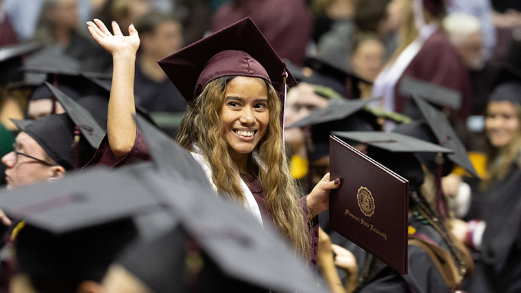 A student at commencement waving and smiling while holding her diploma cover.