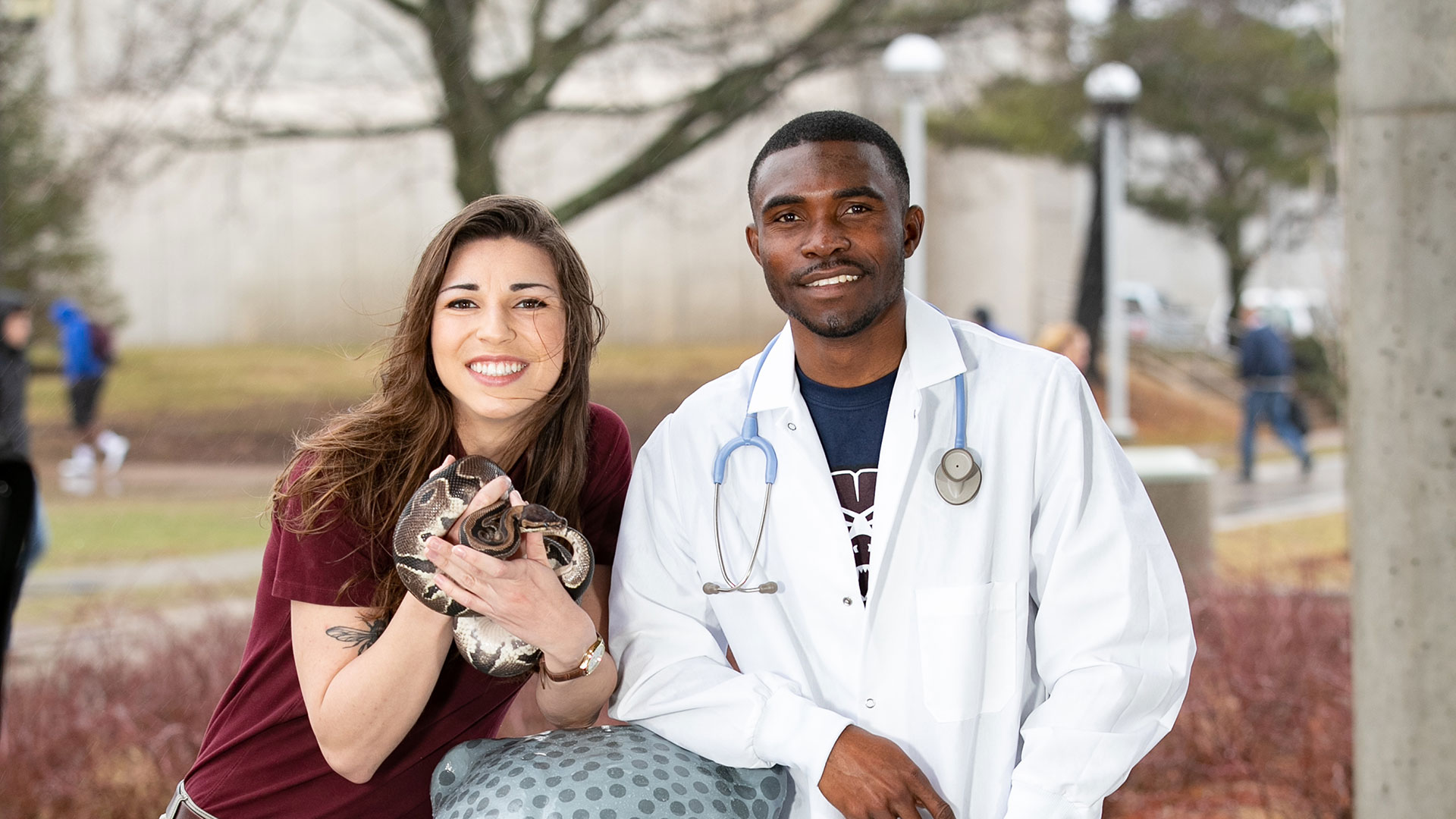 Two biology students pose together outside of Temple Hall. One student is holding a snake, and the other student is wearing a lab coat and stethoscope.