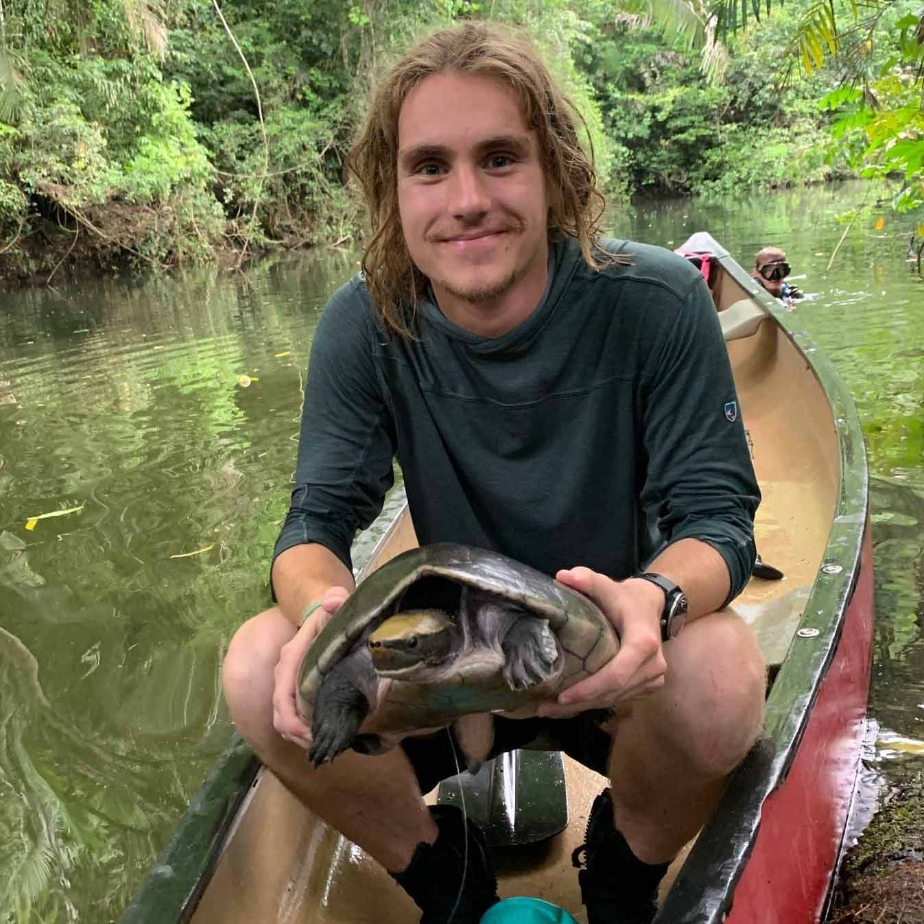A biology student crouching in a canoe while holding up a turtle he caught during field research.