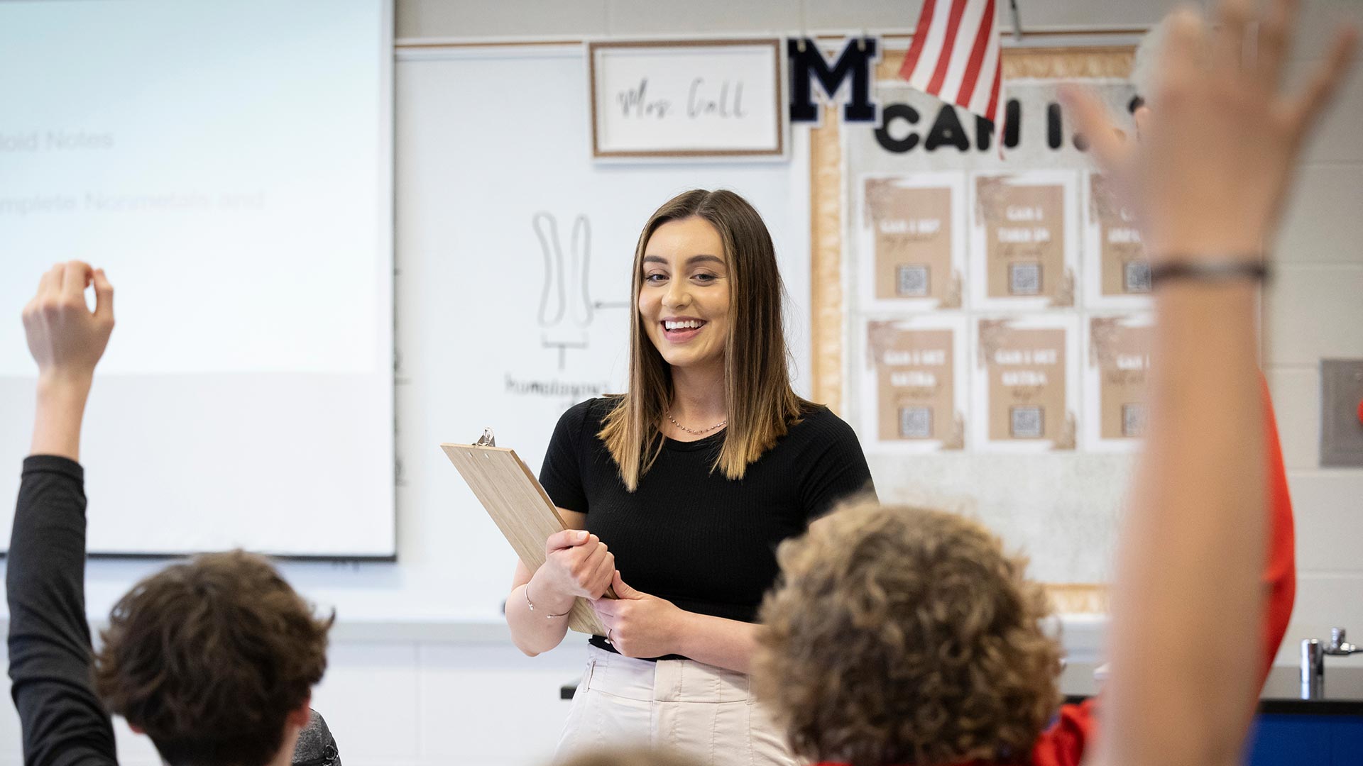 A high school science teacher smiles as her students raise their hands to answer a question during class.