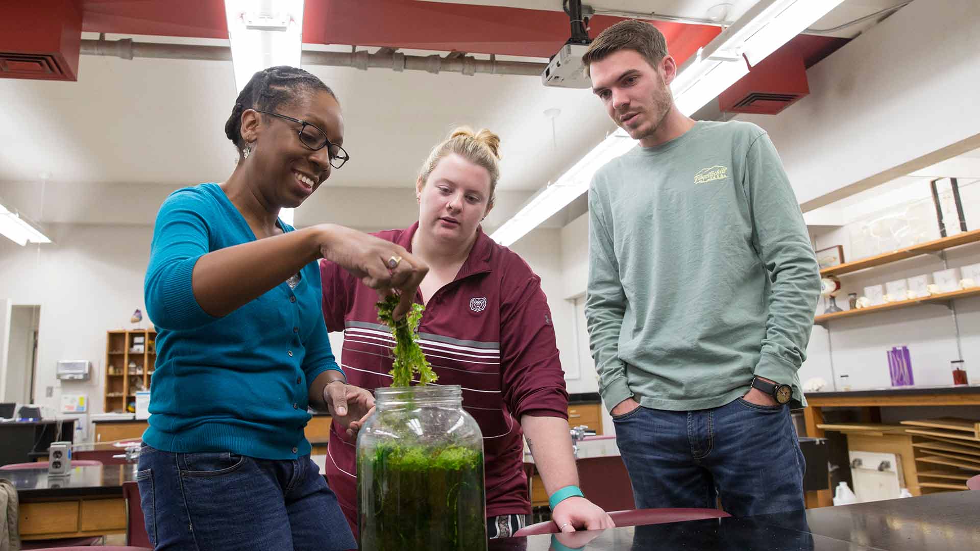 Dr. LaToya Kissoon-Charles pulls out duckweed from a container as two students watch.