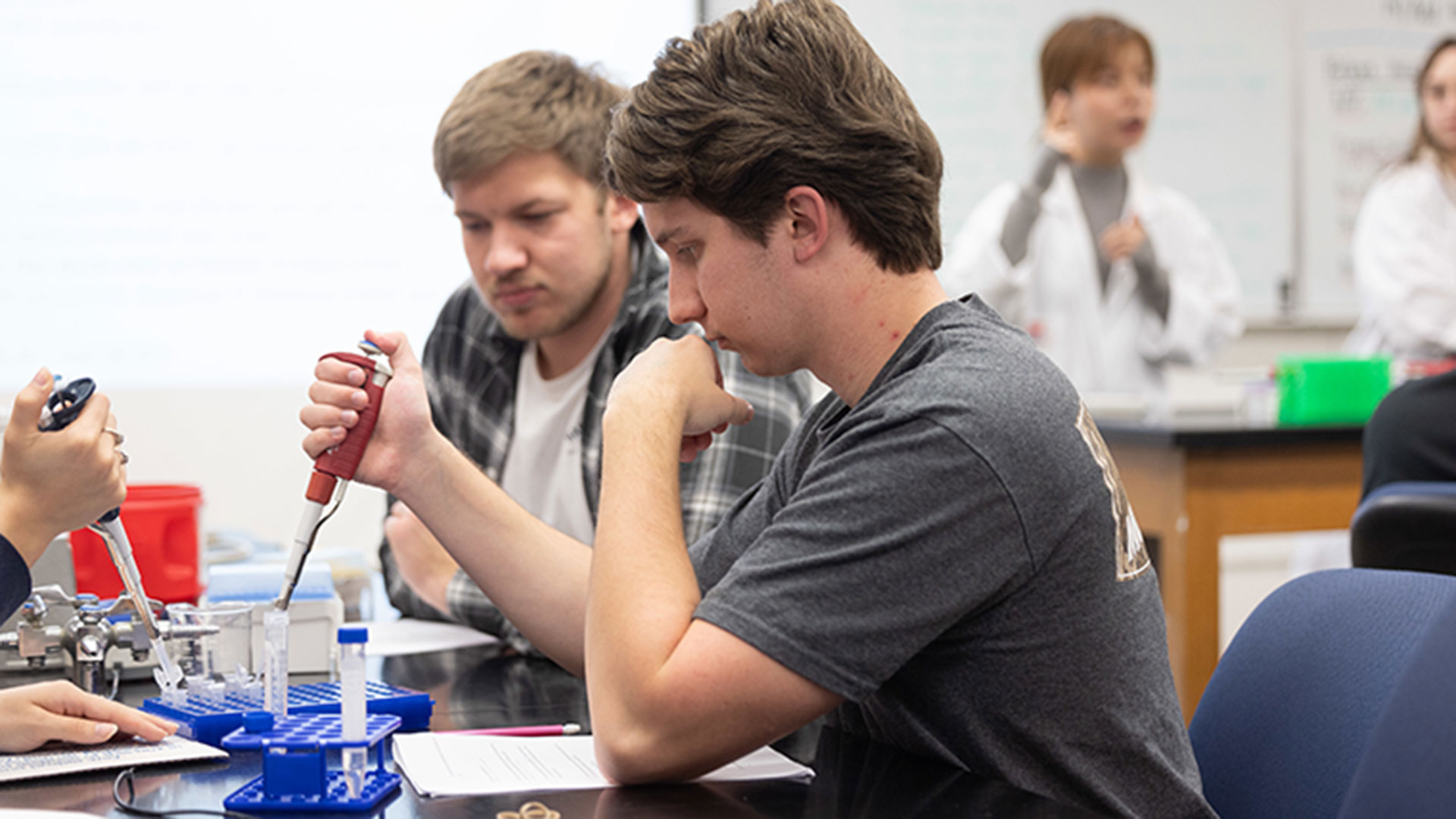 A student uses a pipette to add a liquid to a test tube in his cellular biology class. Beside him, another student watches.