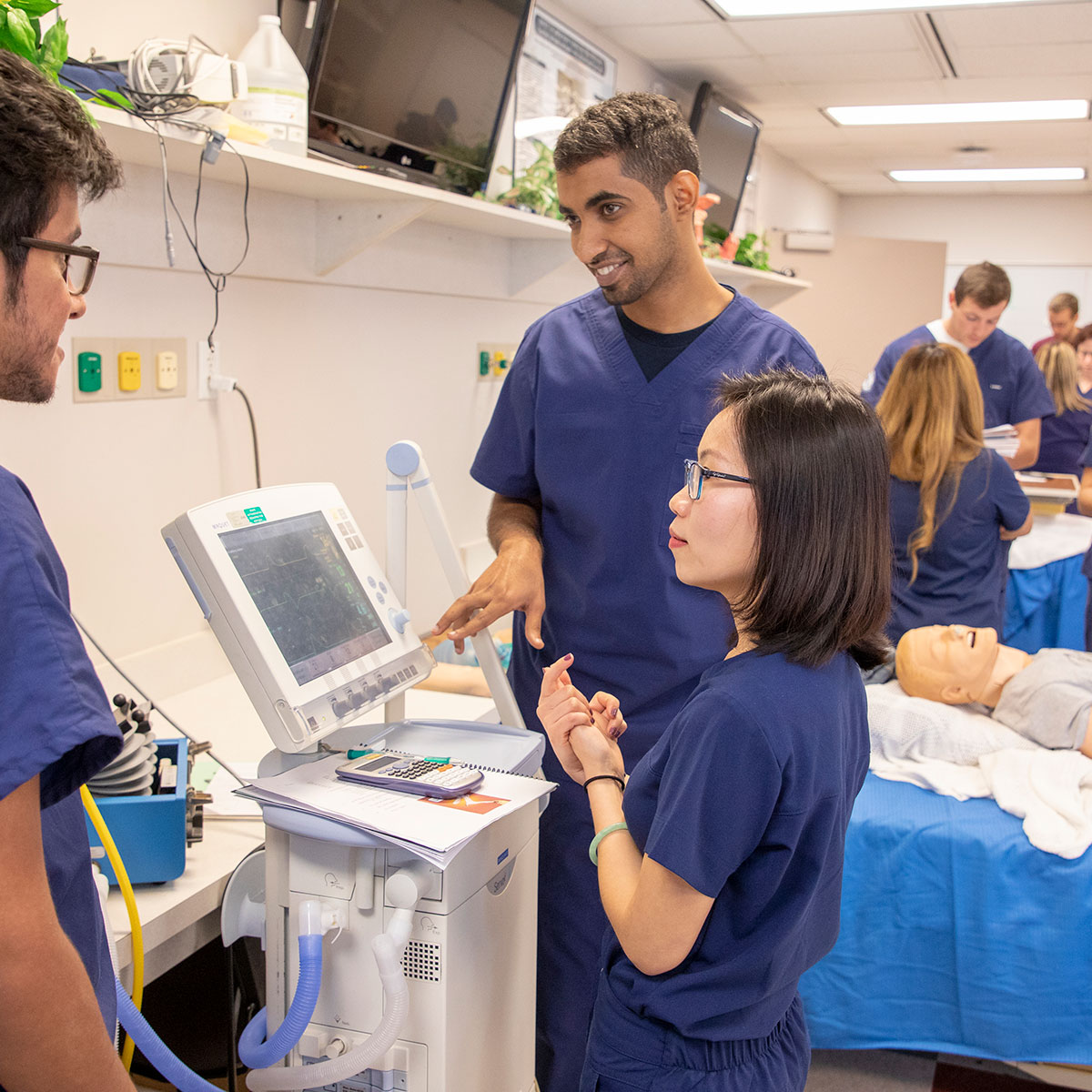 Three medical students read the vitals of a mannequin patient during a practice session.