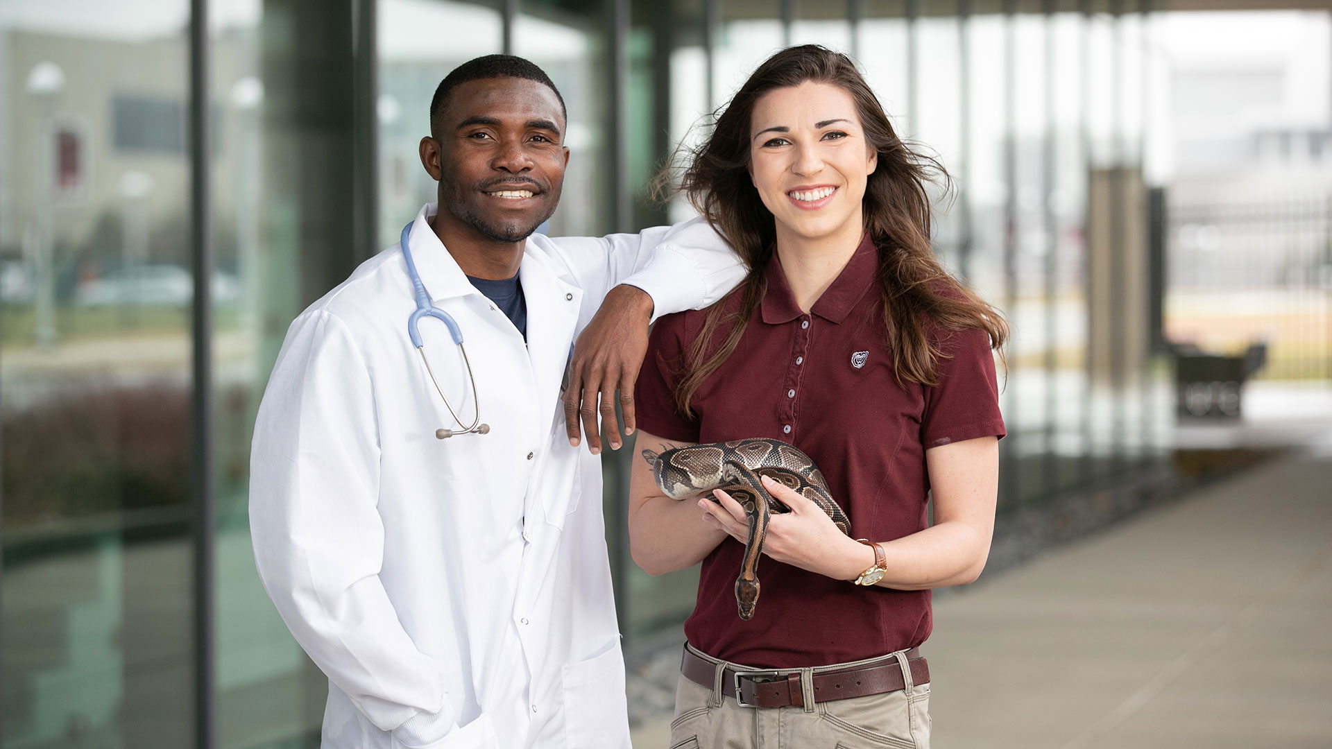 A student in a lab coat standing next to a student holding a snake.