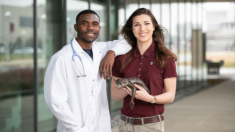 A student wearing a lab coat stands next to a student holding a snake.