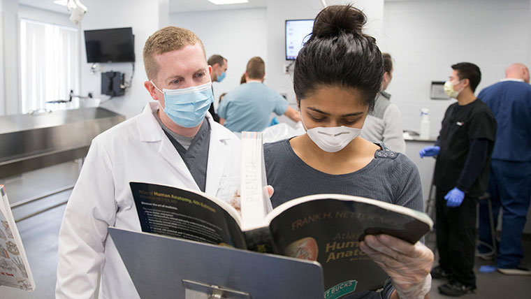 Two physician assistant students refer to an anatomy textbook during a practice activity.