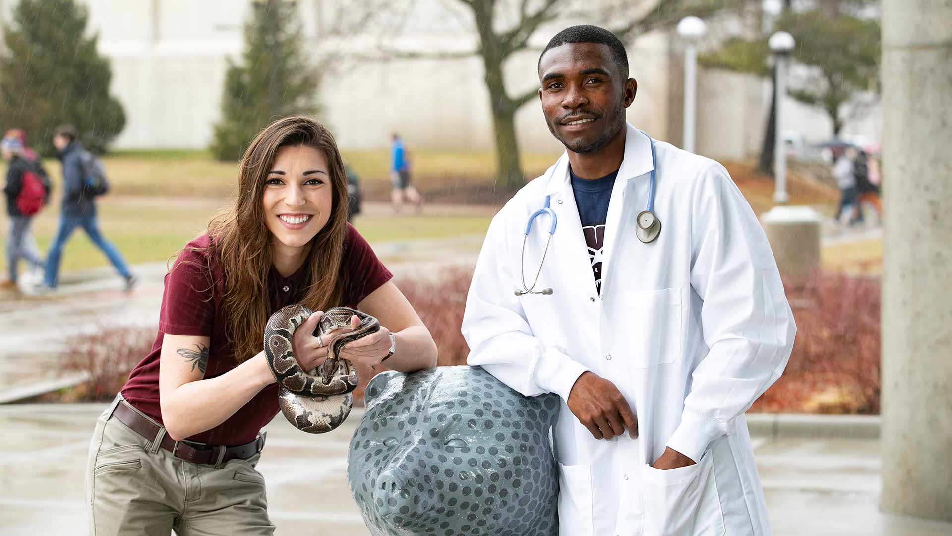Two biology students pose next to a Bear statute outside Blunt Hall. One student is holding a snake. The other student is wearing a white lab coat.