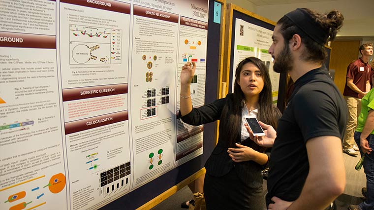A biology student at her posterboard talks about her research topic to another student during Undergraduate Research Day.